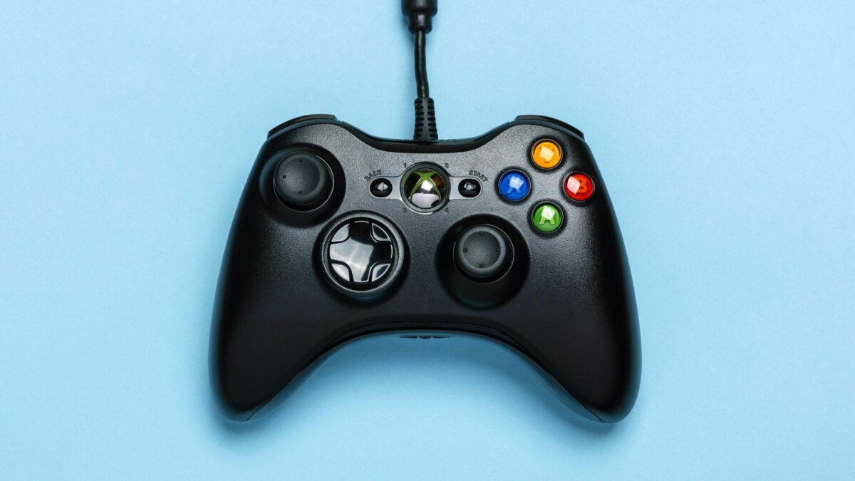 Close-up of a black gaming controller with colorful buttons and a joystick, set against a light blue background.