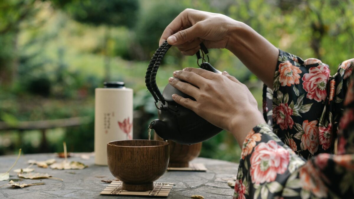 Person in a floral robe pouring tea from a traditional teapot into a wooden tea cup on a table, with a natural outdoor background.