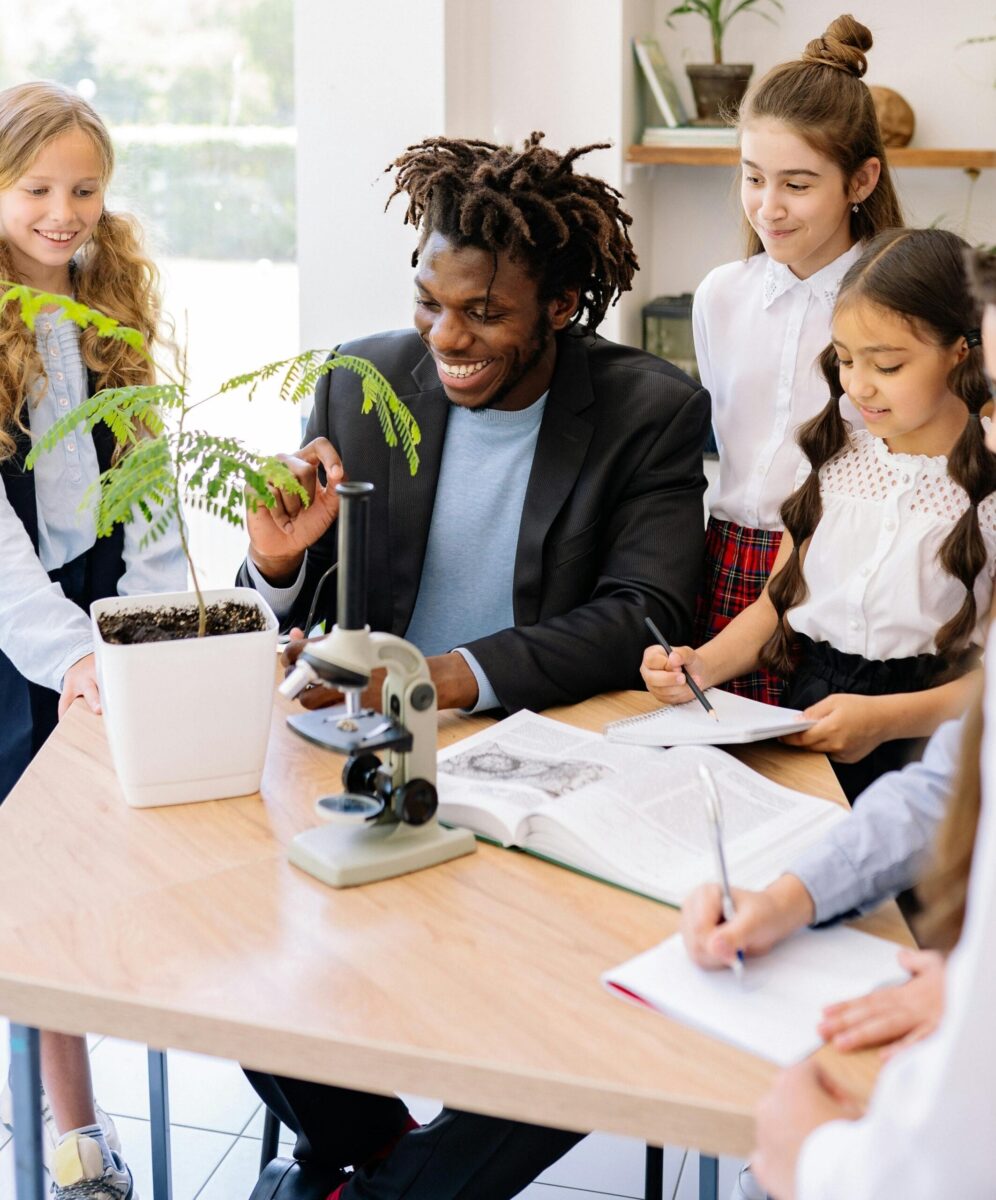 Teacher and a group of four students engaged in a science activity, with a plant and a microscope on the table as they explore and take notes.
