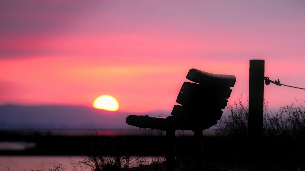 Lonely bench silhouetted against a vibrant sunset, with soft pink and orange hues in the sky and a peaceful landscape.
