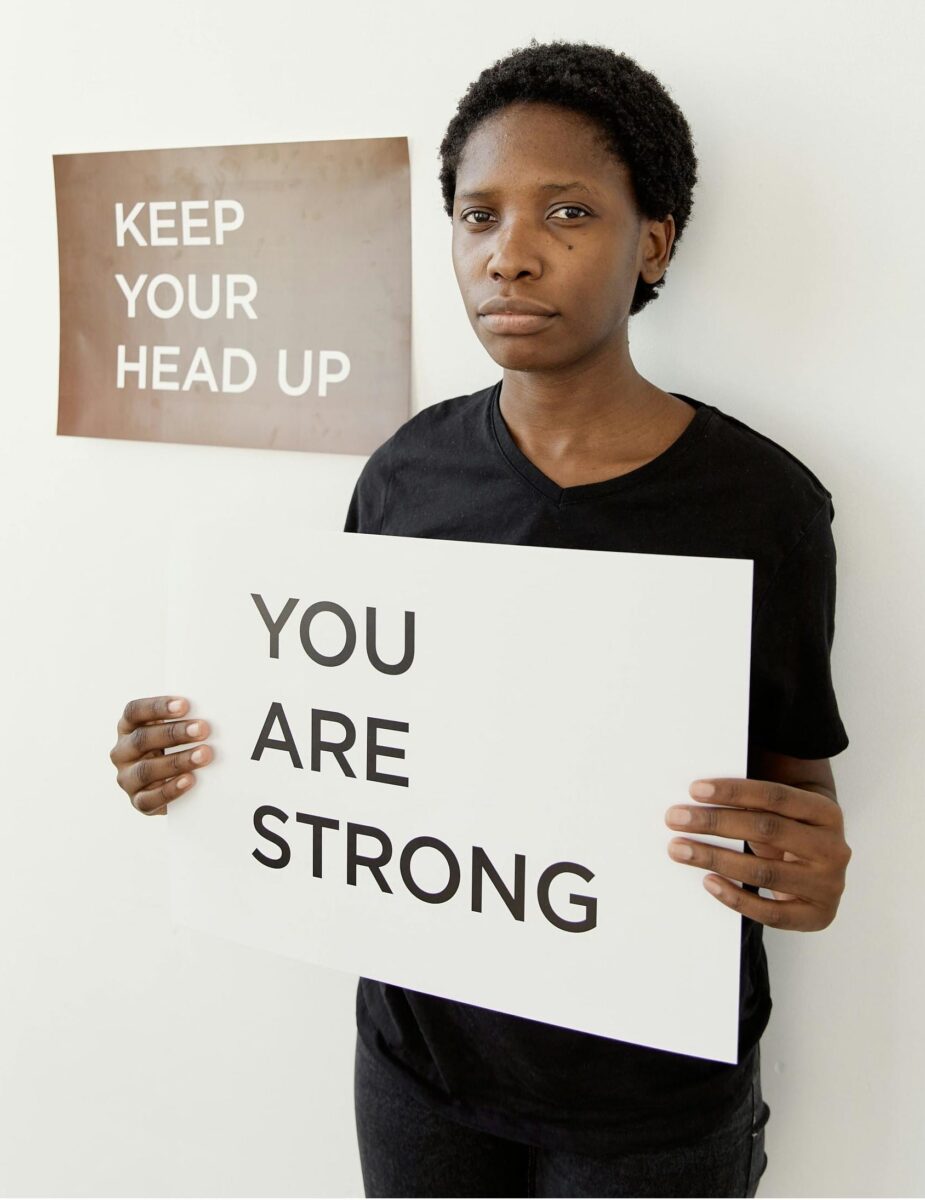 Person holding a sign that reads 'YOU ARE STRONG' while standing under a poster that says 'KEEP YOUR HEAD UP'