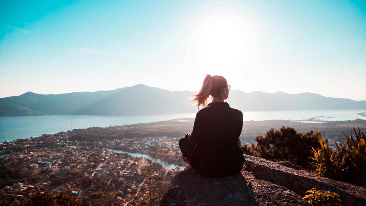 Person sitting on a rock, looking out over a scenic landscape with mountains, water, and a city below, bathed in the warm glow of sunlight
