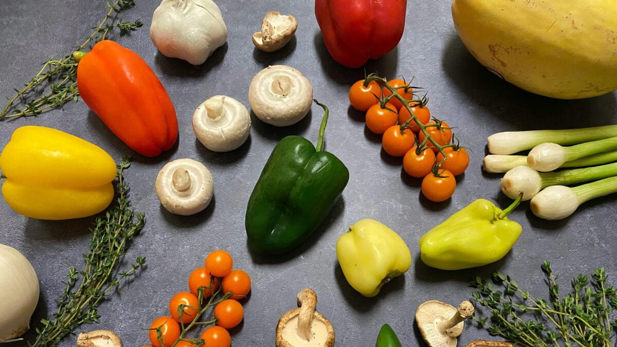 Variety of fresh vegetables arranged on a dark countertop, including bell peppers, mushrooms, tomatoes, garlic, green onions, and thyme.
