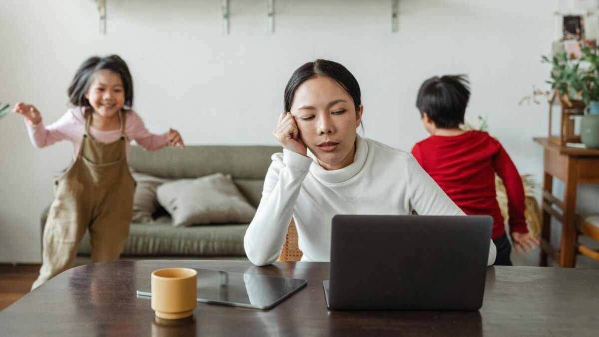 Woman working on a laptop with a tired expression, while two children play in the background, one in a red shirt and the other in overalls, in a cozy home environment.