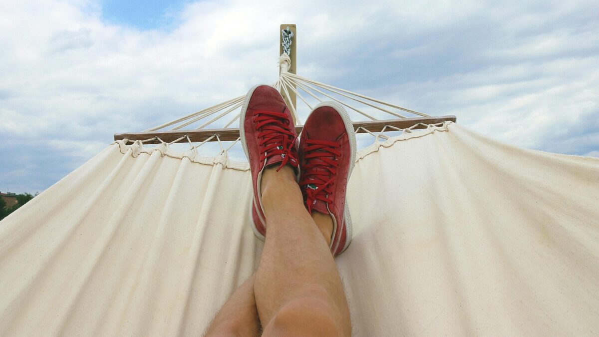 Person relaxing in a hammock, with their legs stretched out and wearing red sneakers, under a partly cloudy sky