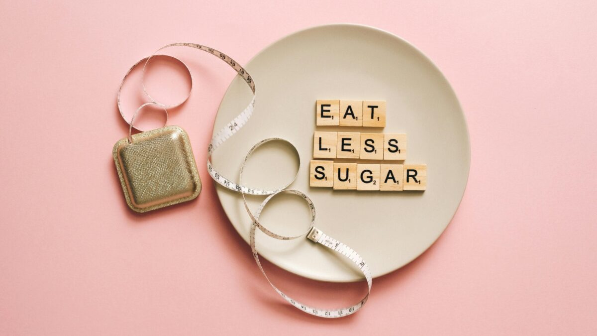 Scrabble tiles arranged on a plate spelling 'EAT LESS SUGAR', with a measuring tape and a tea bag nearby, set on a pink background